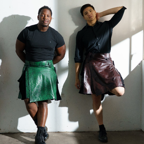 Two people wearing leather kilts against a white wall.