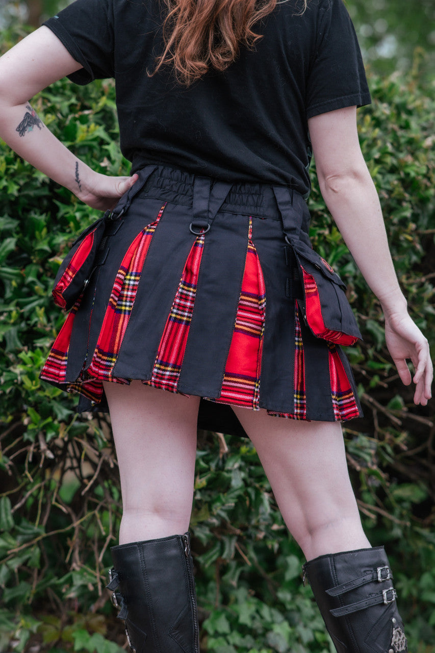 Person wearing a black and red plaid mini kilt with a black top and knee-high boots against a green leafy background.