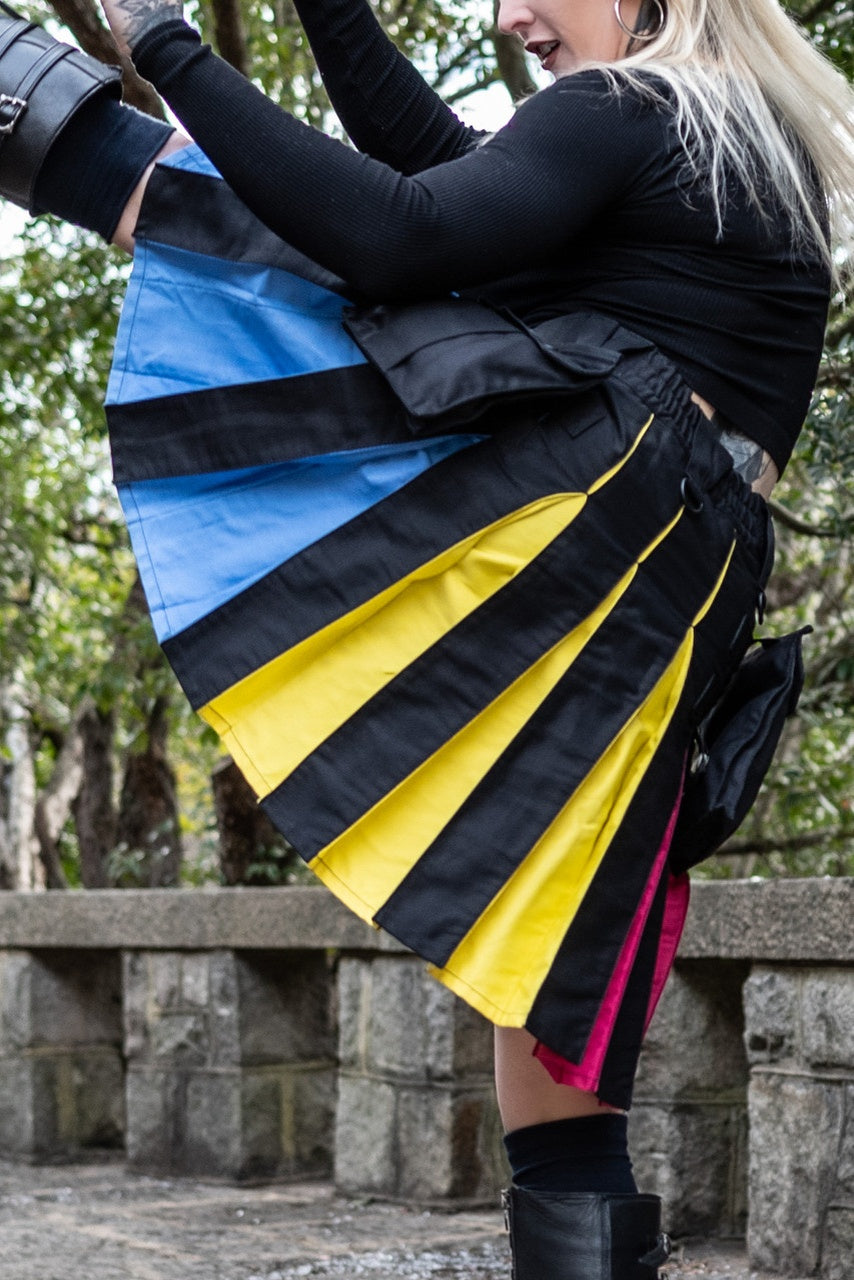 Woman doing a high kick to show off a black kilt with blue, yellow, and pink pleats for pan pride with a blurred background