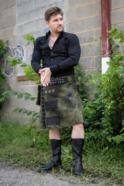 Man wearing a dark green kilt with black accents standing against a gray brick wall.