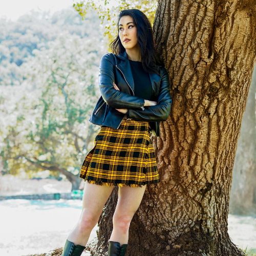 Woman in a blue jacket and yellow plaid kilt leaning against a tree in a park.