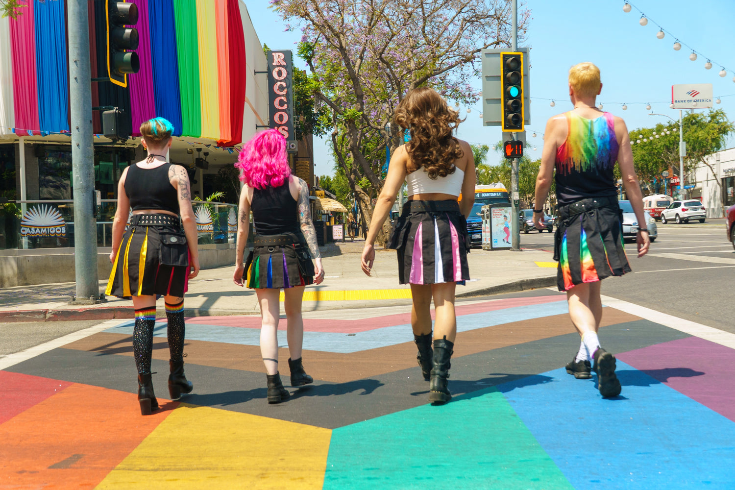 Four people in colorful Pride-themed kilts crossing a rainbow crosswalk on a city street.