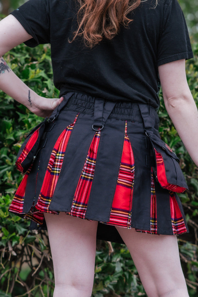 Person wearing a black and red plaid mini kilt with a black top and knee-high boots against a green leafy background.