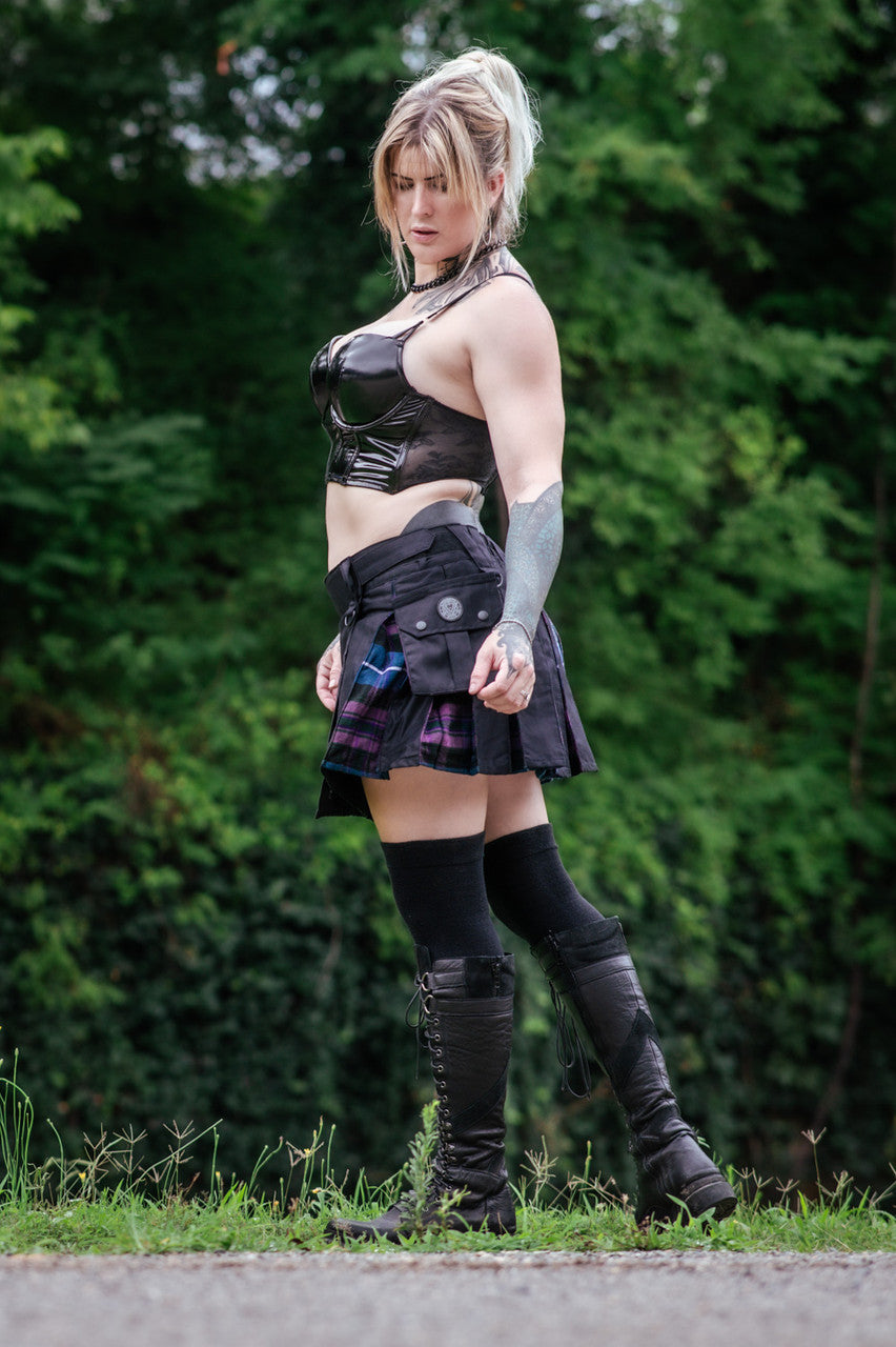Woman in black top wearing a black mini kilt with purple and blue plaid details in a forest-y setting