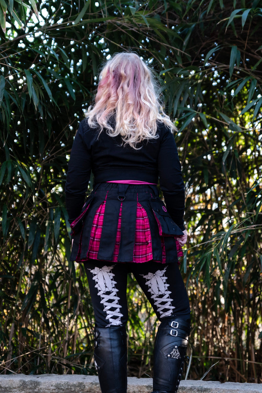 Person wearing a black and pink plaid mini kilt and leather boots, standing in front of a bamboo background.