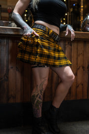 Person wearing a yellow plaid mini kilt with tattoos visible on arms and legs, standing against a wooden background.