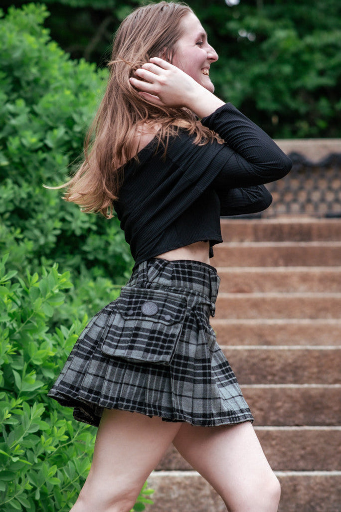Woman in a grey plaid mini kilt and black top standing on steps with greenery in the background