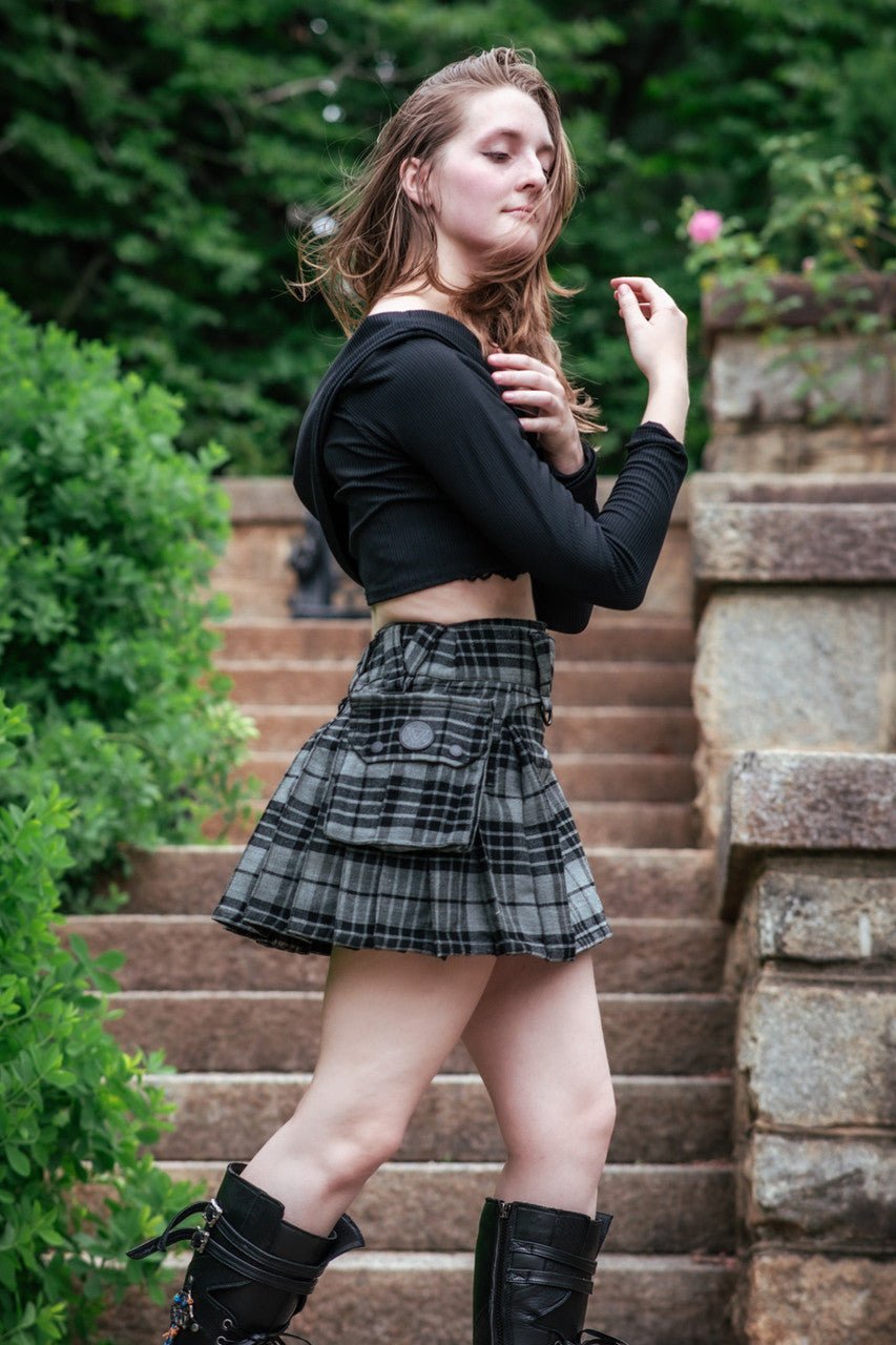 A woman wearing a black top and grey plaid mini kilt stands on stone steps outdoors.