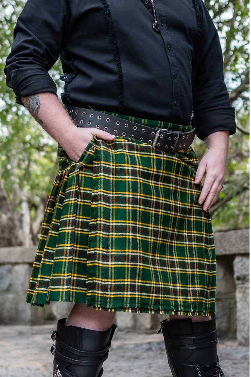 Person wearing a green and yellow plaid kilt with a black shirt and boots outdoors.