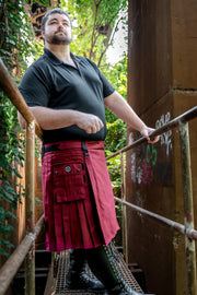 Man wearing a red kilt standing on a metal staircase with greenery in the background