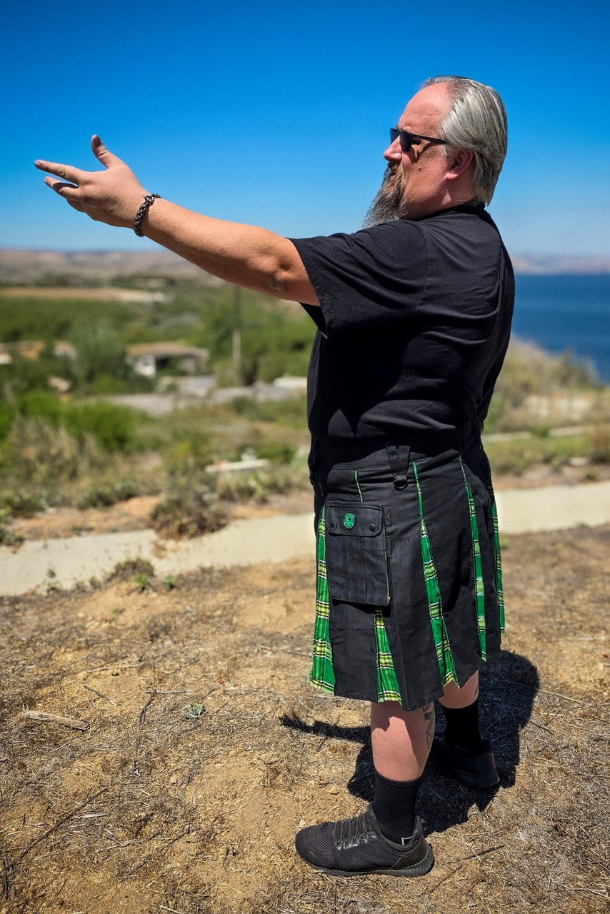Man wearing a black kilt with green accents standing outdoors with a scenic background.