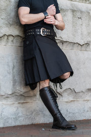 Person wearing a black kilt and boots against a stone wall.