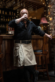 Person standing in a bar, holding a drink, in a khaki kilt and black button-up.