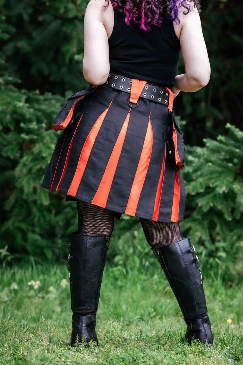 Person wearing a black and orange pleated kilt with black knee-high boots in an outdoor setting.