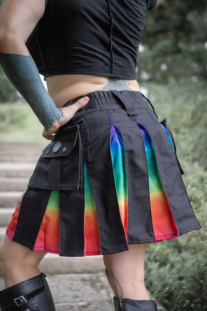 Back view of a person wearing a black, knee-length kilt with ombre rainbow accents outdoors.
