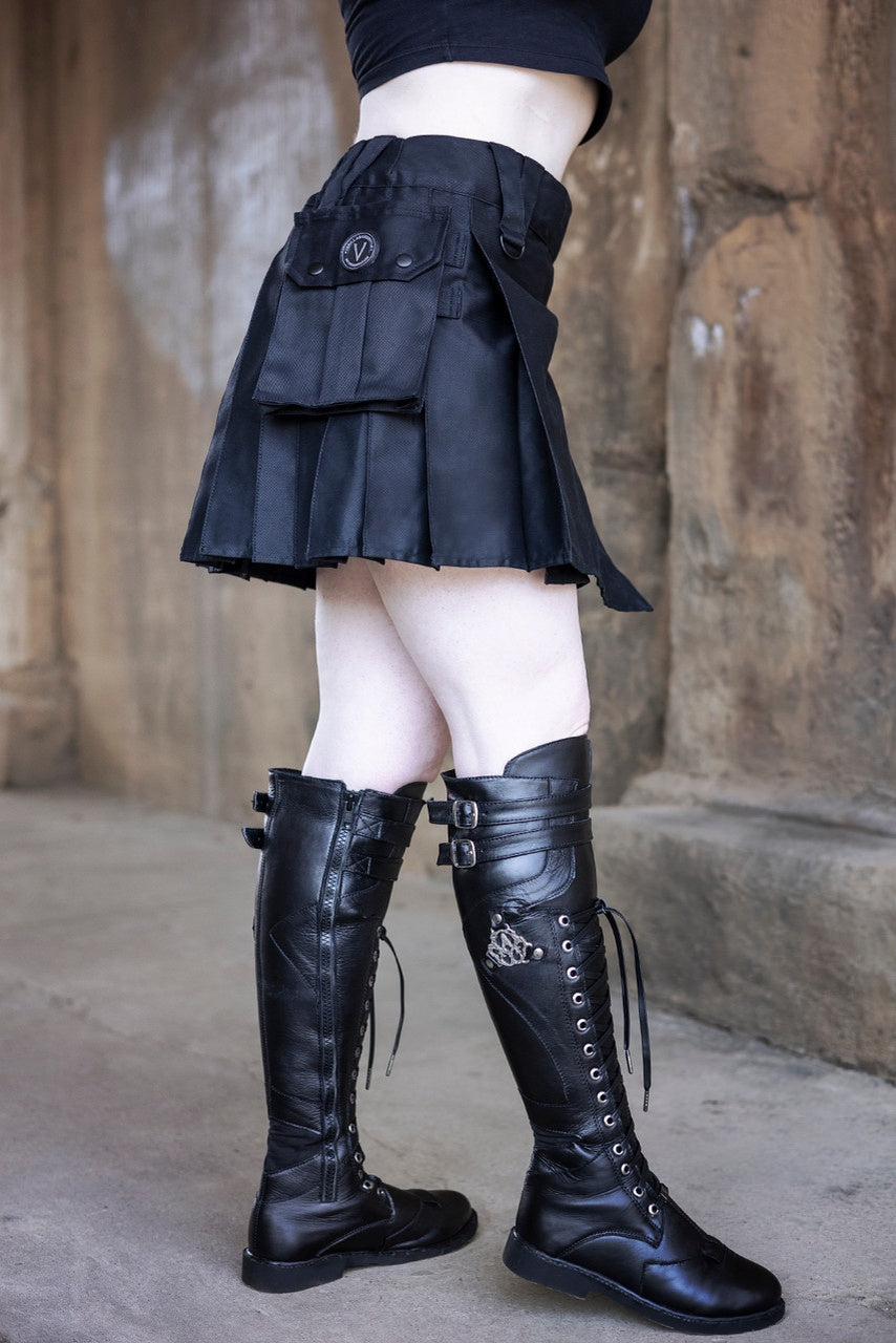 Person wearing a black mini kilt and high black boots against a stone wall.