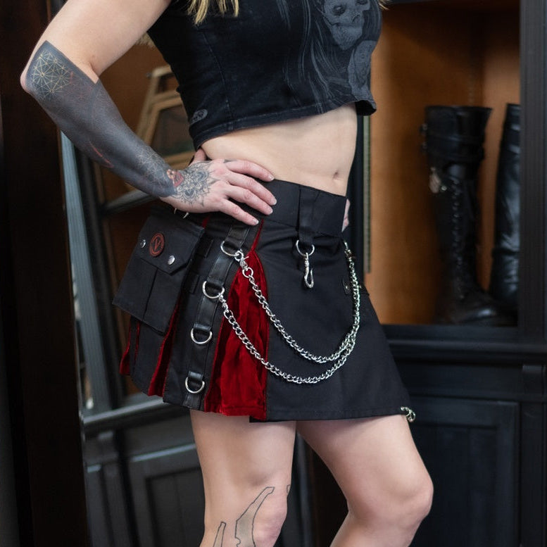 Person wearing a black top and a black short kilt with red velvet pleats and boots in a room setting.
