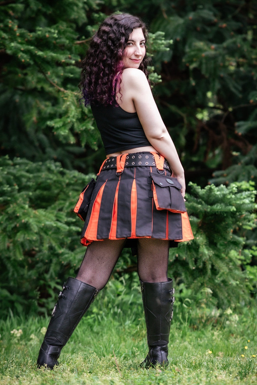 Person wearing a black and orange pleated kilt with a black top and black boots outdoors.