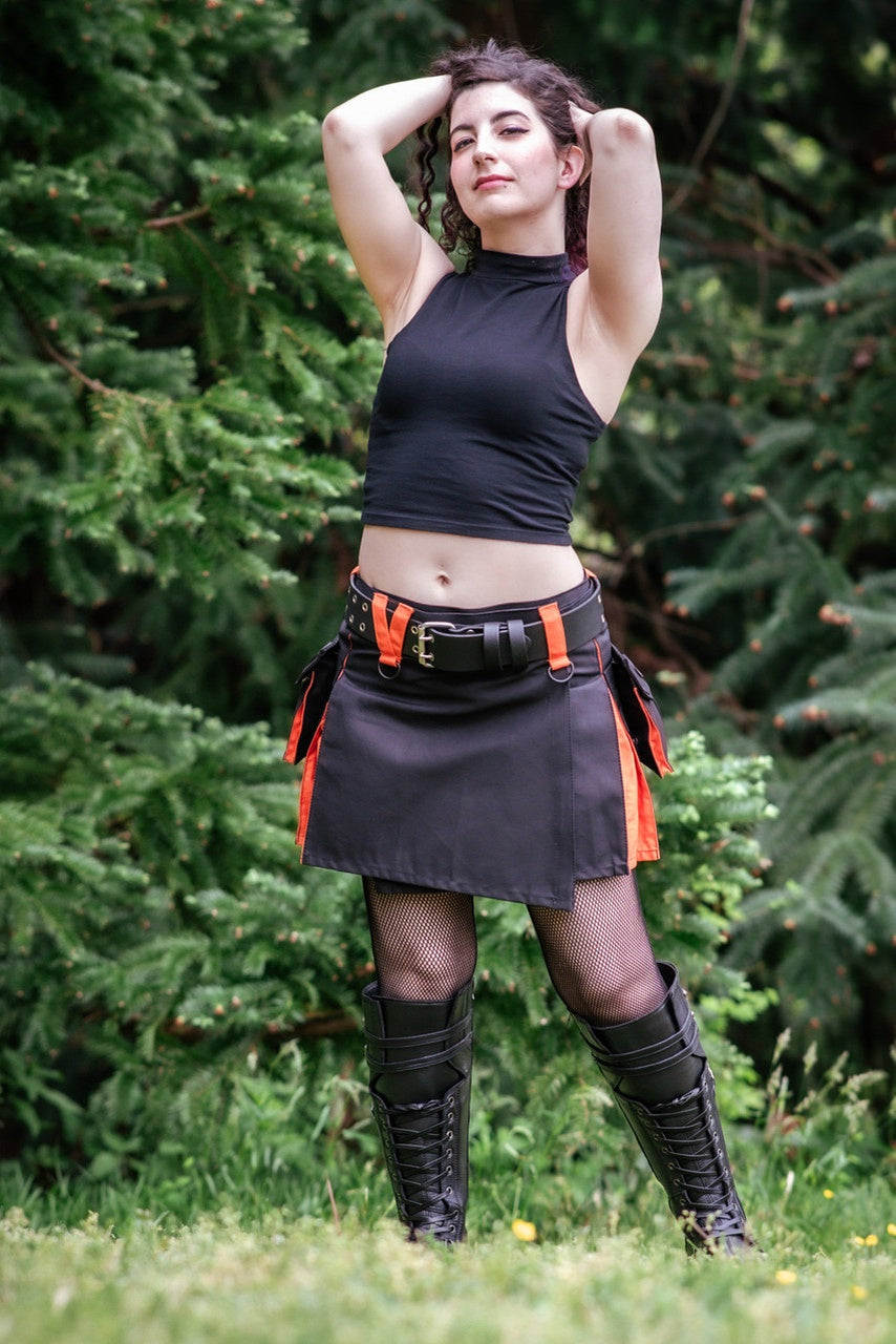 Woman in black top and kilt with orange accents standing outdoors among green trees.