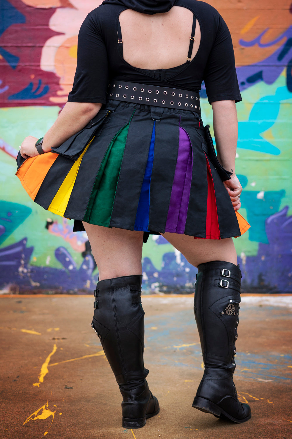 A person wearing a black outfit with a colorful kilt in front of a graffiti wall.