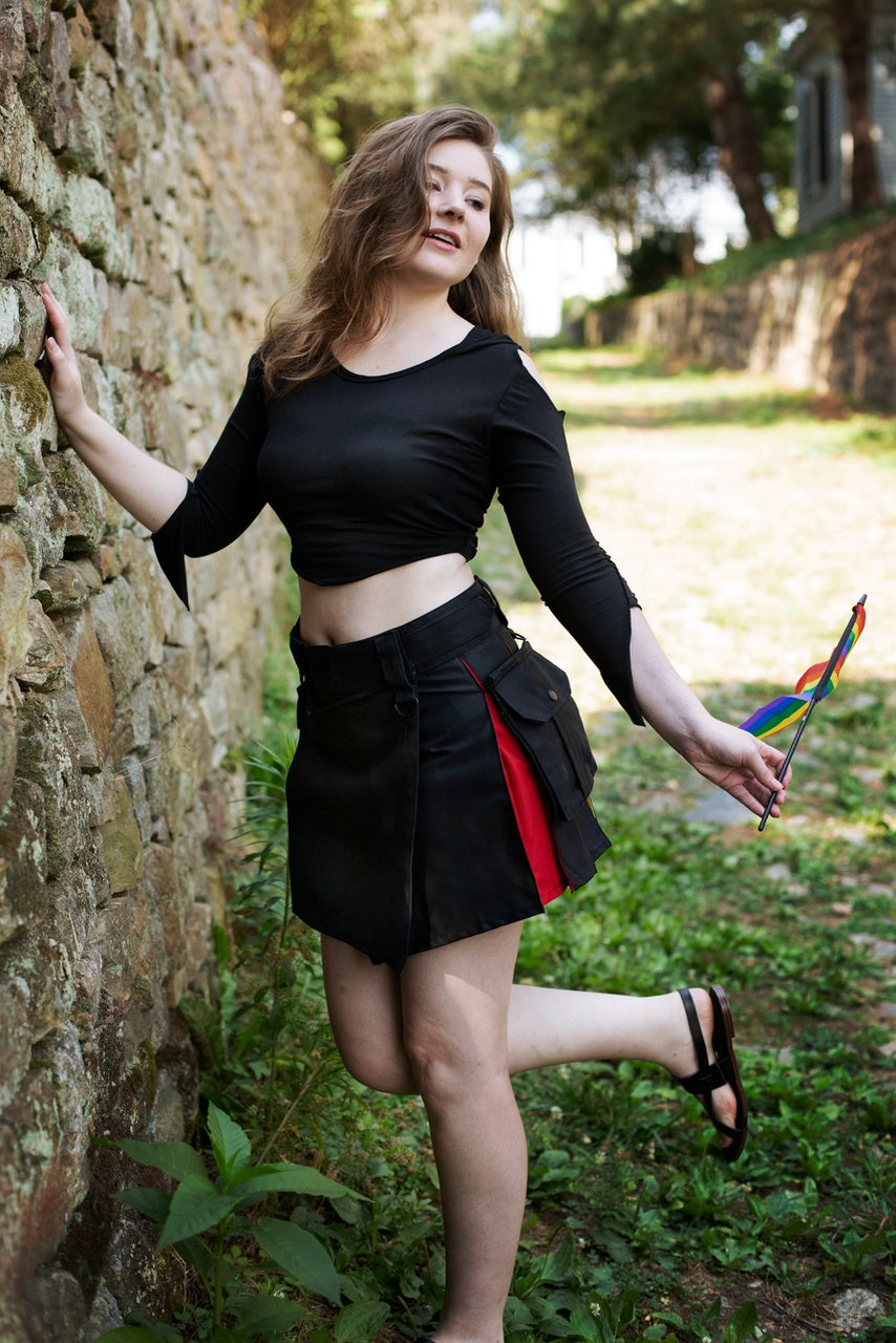 Woman in black top and short black and red kilt standing against a stone wall outdoors