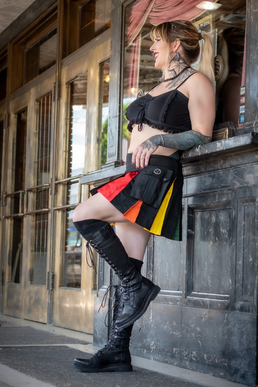 A woman wearing a black mini kilt with rainbow pleats, leaning against a building.
