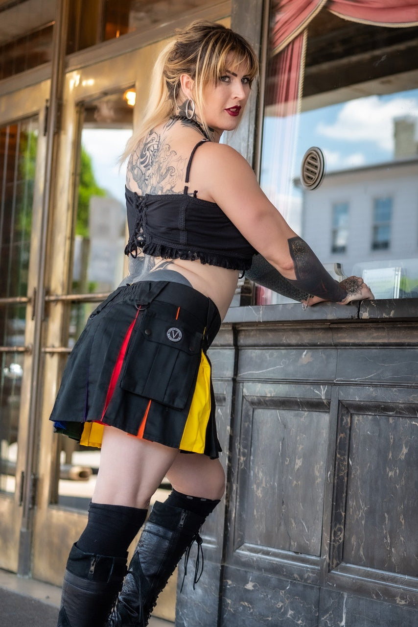 Woman in a stylish outfit with a black and rainbow mini kilt in front of a building entrance.