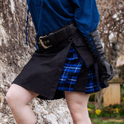 Person in a blue top and plaid kilt leaning against a tree in an outdoor setting