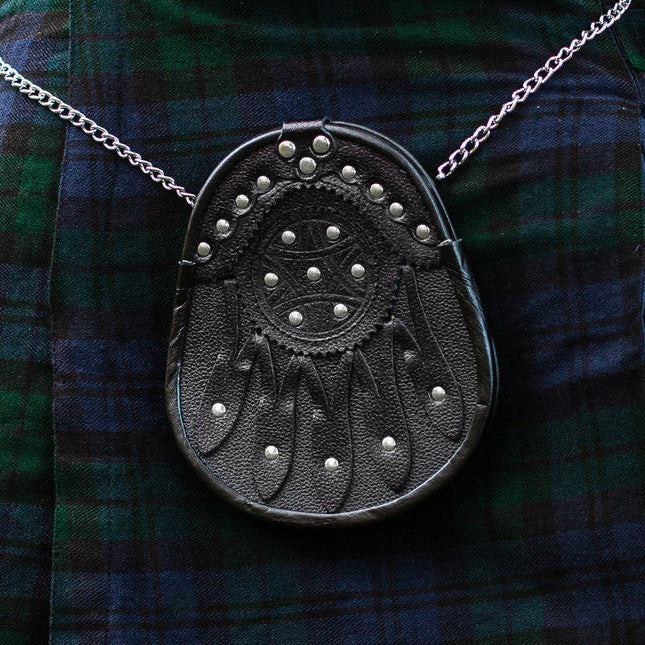 Close up of a black leather sporran with leather detailing and silver studs hanging at the front of a plaid kilt.