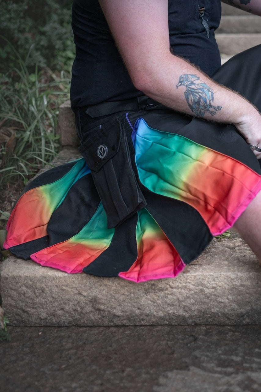Person wearing a black kilt with an ombre rainbow pattern on a stone step.