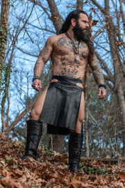 Man with tattoos and a beard wearing a leather kilt in a forest setting