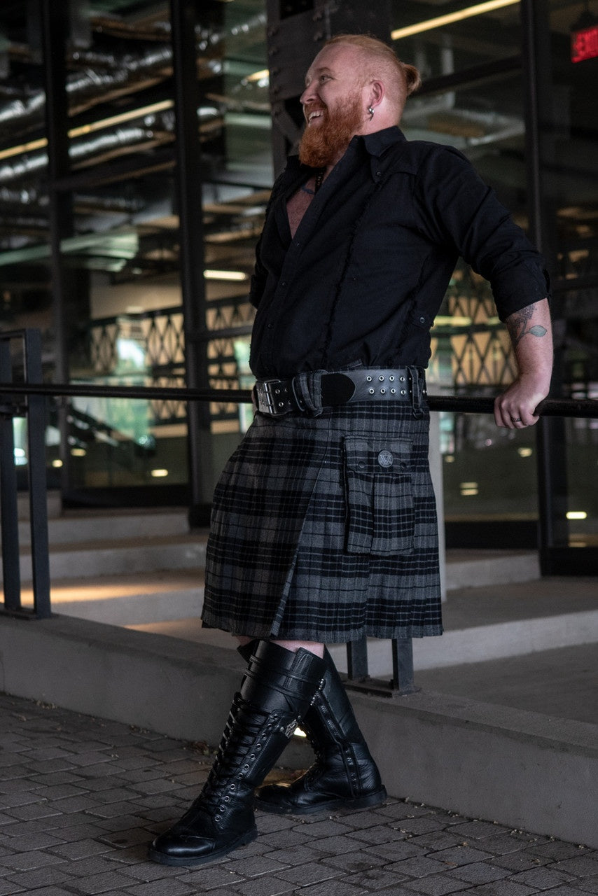 Man wearing a black shirt, plaid kilt, and black boots standing in front of a modern building.