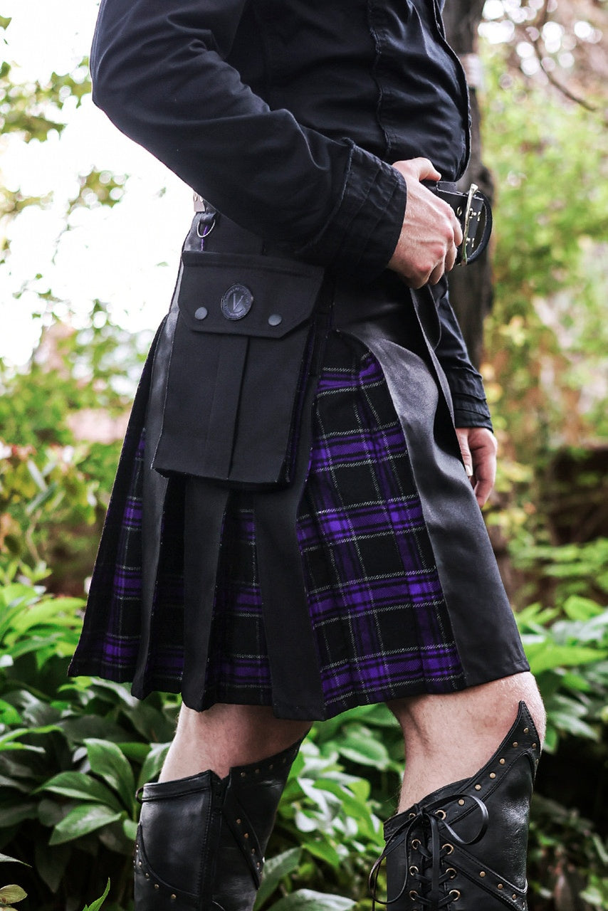Person wearing a black coat and plaid kilt with greenery in the background