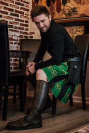 Man wearing a green and black plaid kilt sitting on a chair in a room with a brick wall.