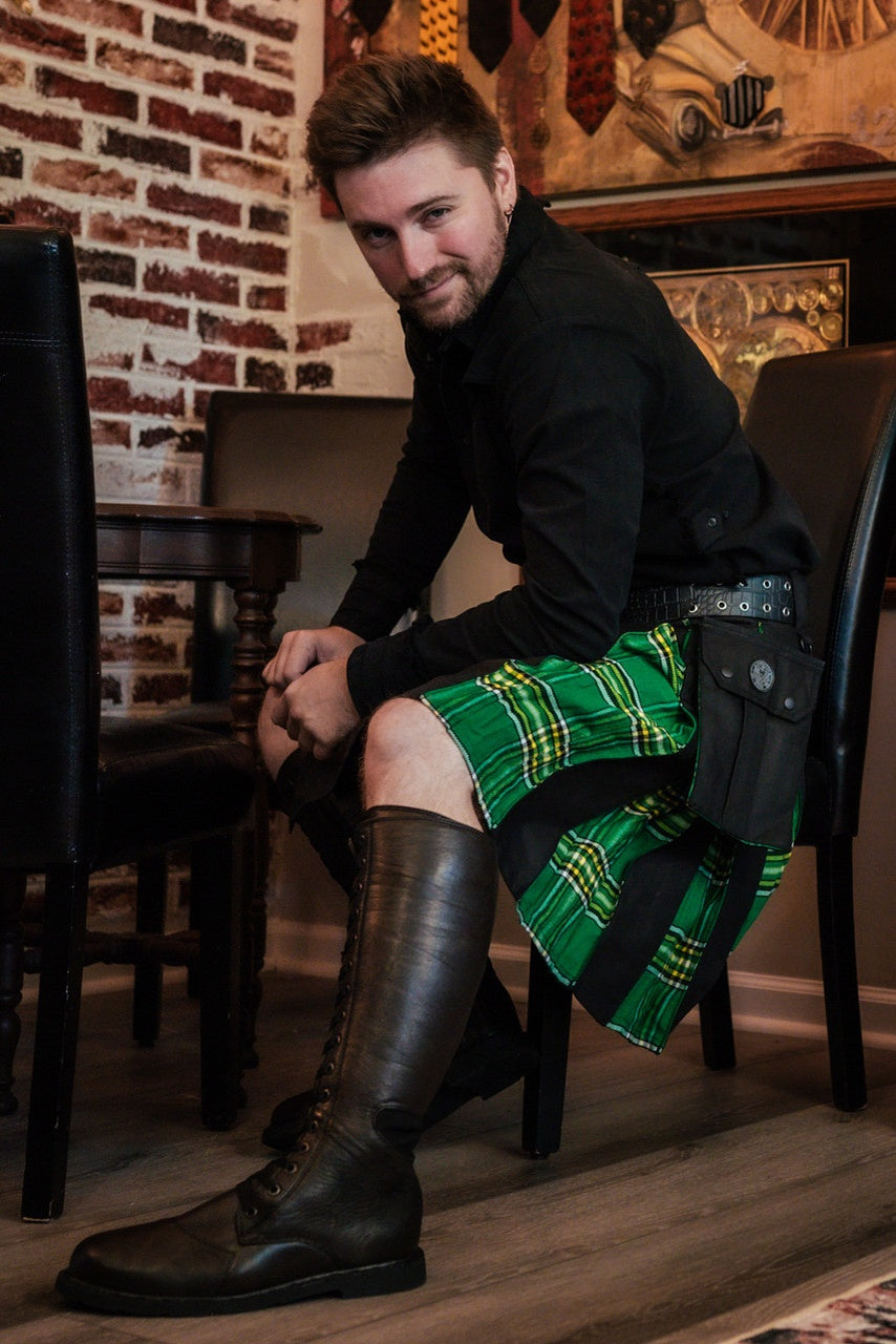 Man wearing a green and black plaid kilt sitting on a chair in a room with a brick wall.