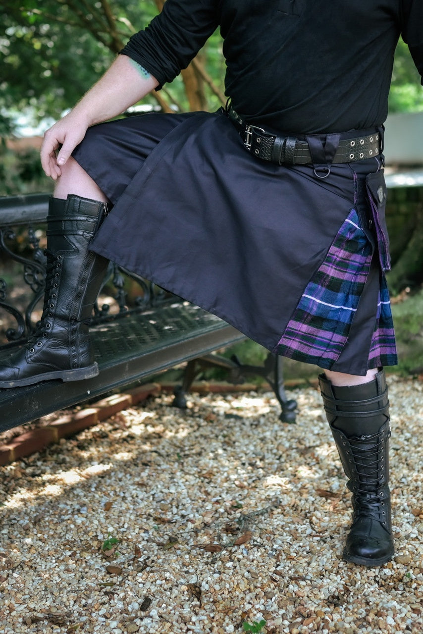 Person wearing a dark kilt with a plaid accent, black boots, and a black top outdoors.
