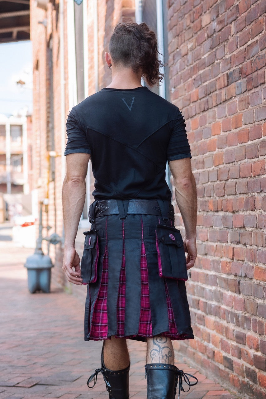 Person wearing a black shirt and plaid kilt standing against a brick wall.