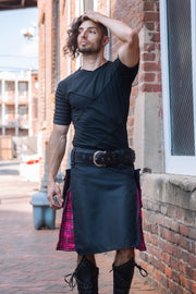 Man wearing a black kilt with a pink plaid lining, standing against a brick wall.