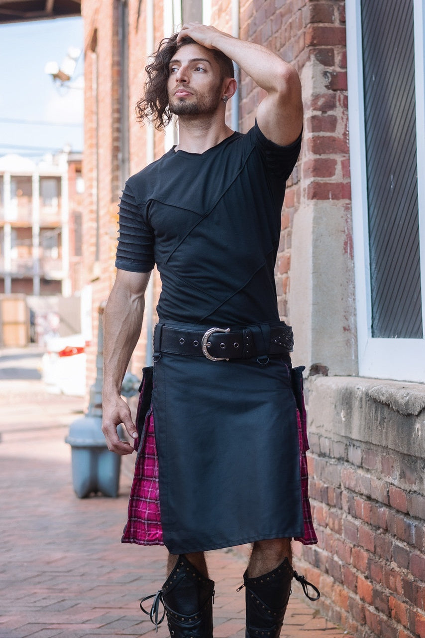 Man wearing a black kilt with a pink plaid lining, standing against a brick wall.