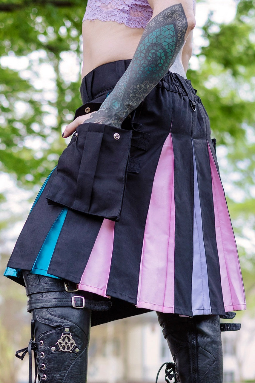 Person wearing a black kilt with pink, blue, and white pleats for trans pride with tattoos on arms, standing outdoors.