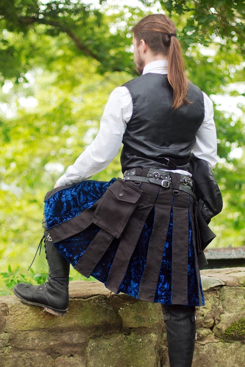 Person wearing a black kilt with blue velvet lining, standing outdoors with greenery in the background.