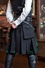 Person wearing a black kilt with green velvet accents and a white shirt, standing indoors.