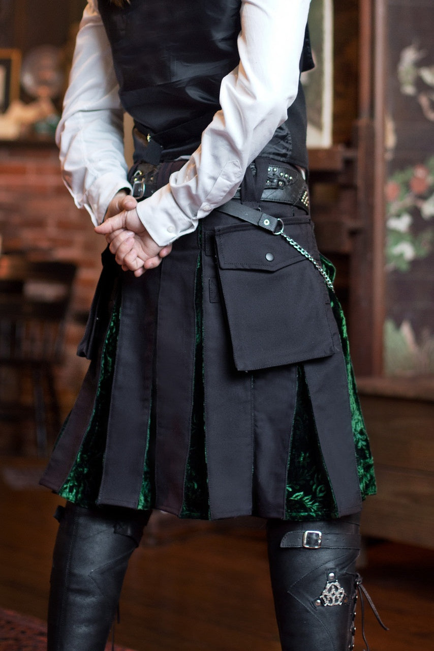 Person wearing a black kilt with green velvet accents and a white shirt, standing indoors.