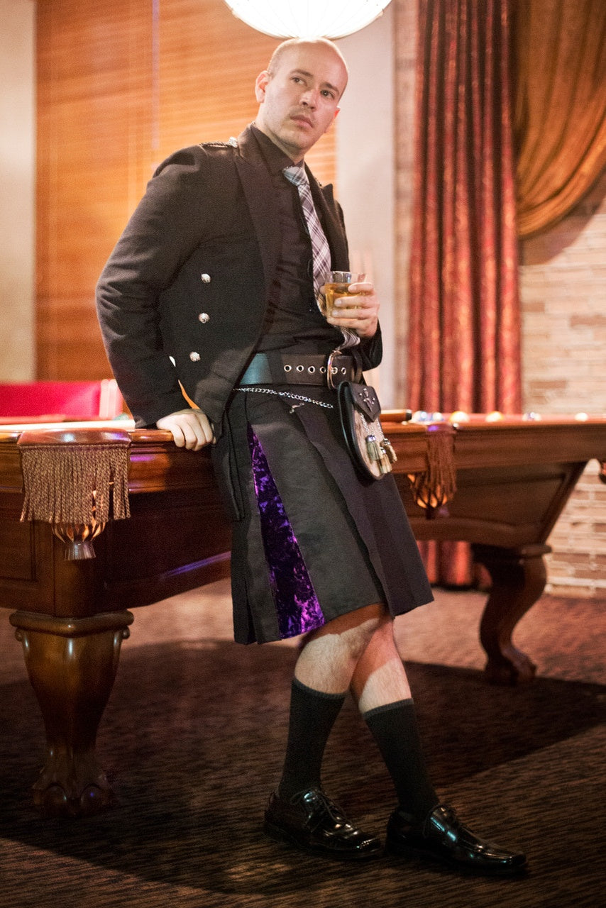 Man in a black and purple velvet kilt holding a glass of whiskey in a room with a pool table and red curtains.