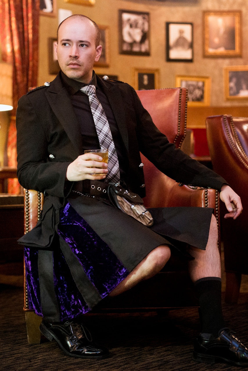 Man in a black jacket and black and purple velvet kilt sitting in a chair holding a drink.