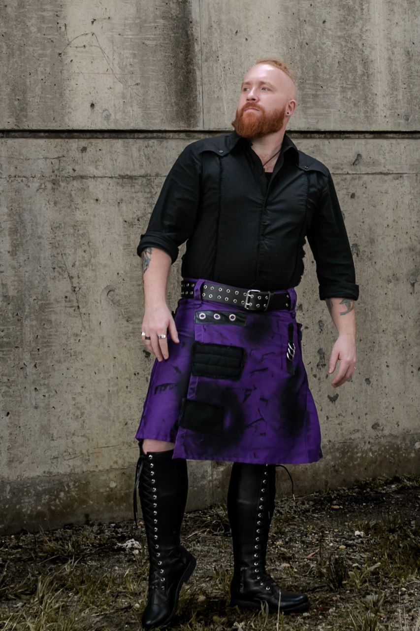 Man wearing a black shirt, purple kilt, and black boots against a concrete wall.