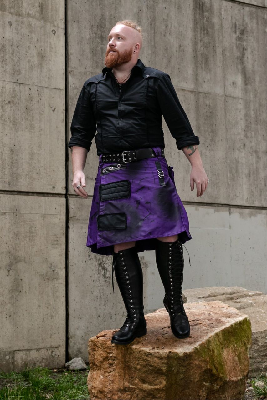 Man wearing a black shirt, purple kilt, and black knee-high boots standing on a rock against a concrete wall.