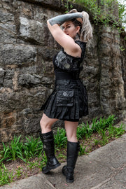Woman in a black outfit with a black camo mini kilt and boots standing against a stone wall.