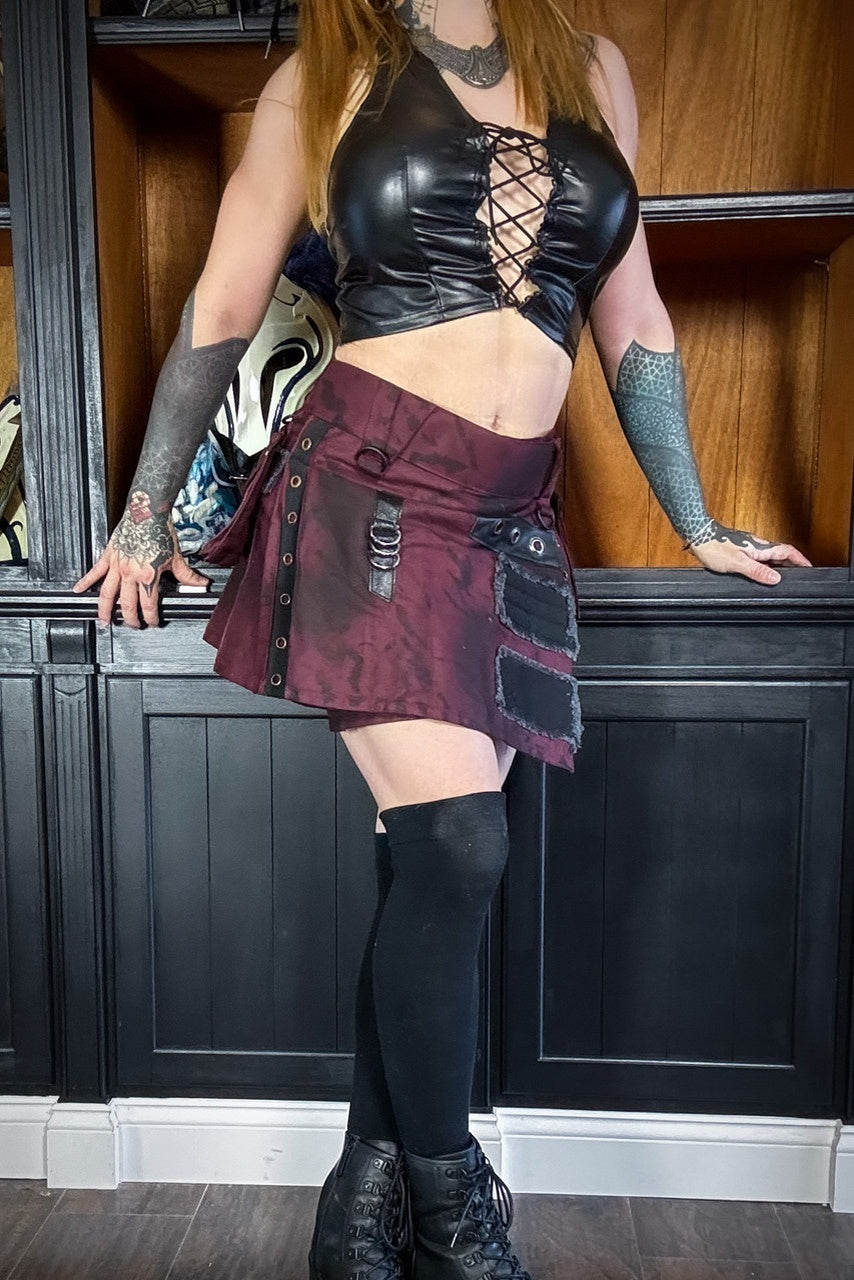 Person wearing a black leather top and burgundy and black mini kilt with black knee-high stockings and boots indoors.