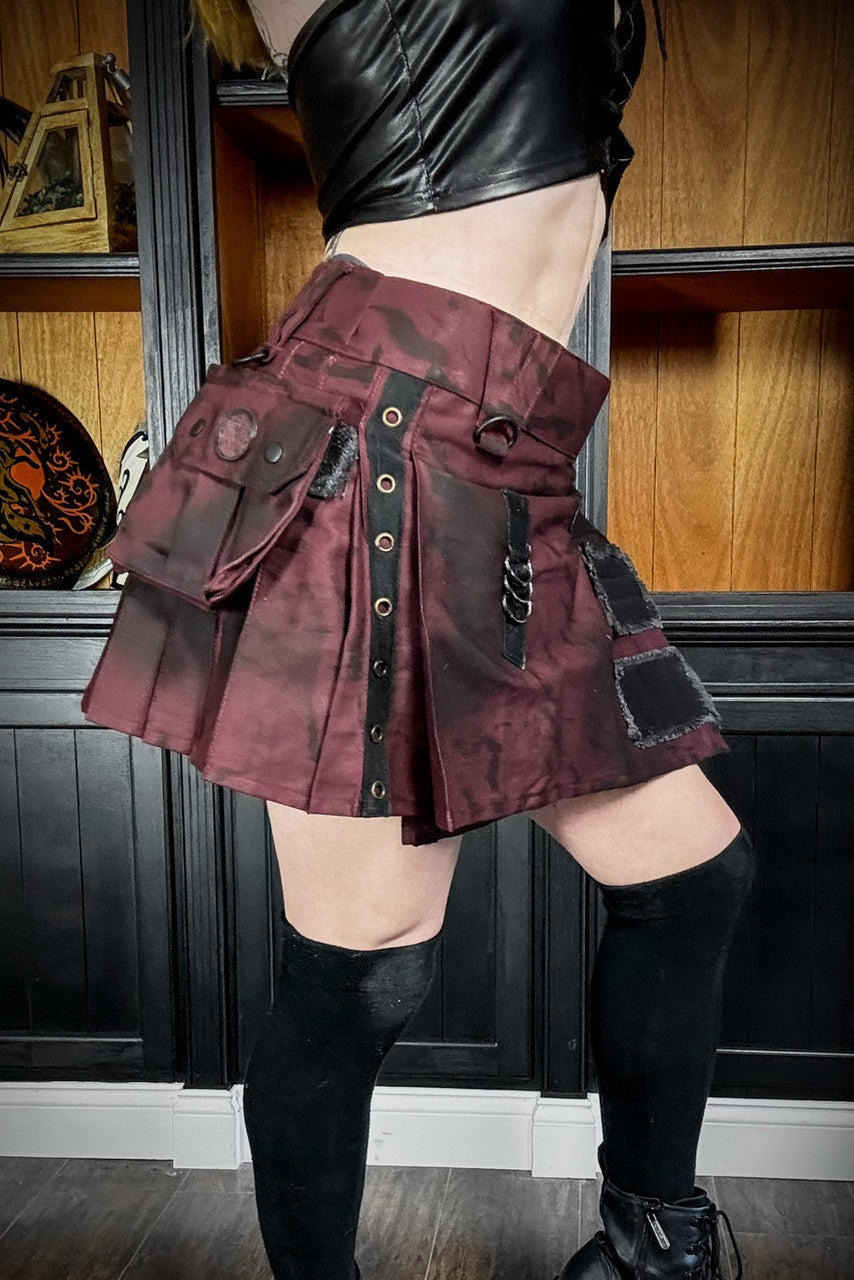 Close up of a person wearing a burgundy pleated mini kilt with black accents in a room with wooden paneling.